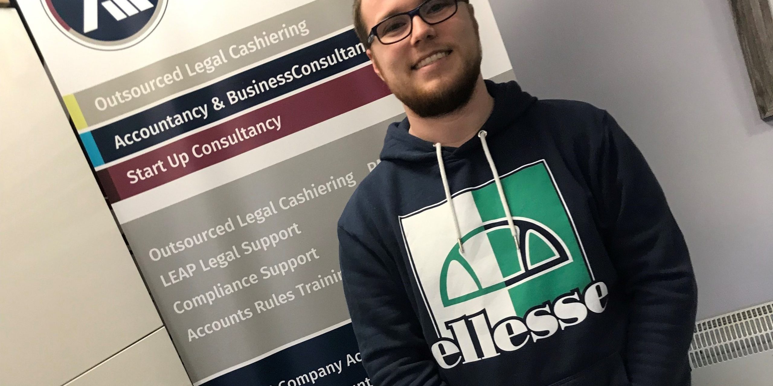 Josh in office Josh in the office standing in front of an Ascentant roller banner