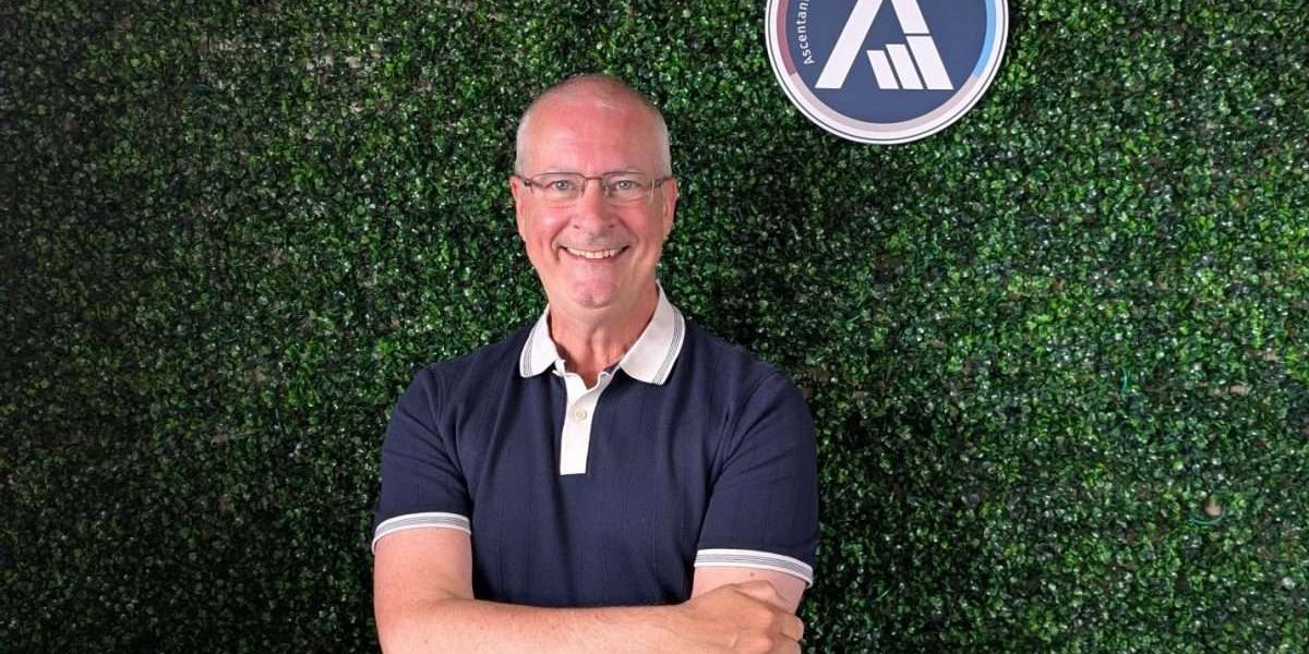 Adrian smiling at the camera with a faux green wall and Ascentant logo behind him