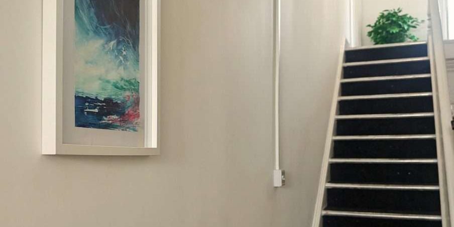 A photo of the artwork hung on the left hand wall with the stairs leading the way up to the first floor in the background.