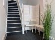 Photo of an area at the bottom of the dark grey carpeted stairs - a few white chairs and a green plant on the floor.